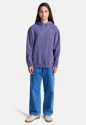Lila Baumwoll-Hoodie mit Vordertasche und gesticktem Logo, kombiniert mit blauen Jeans mit weitem Bein und hellen Sneakers. Einfaches Design, lockere Passform.