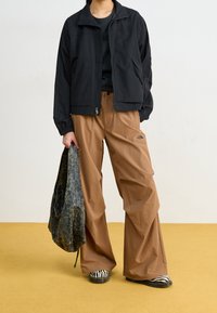 Svart zip-up jacka, bruna vida byxor med logo, randiga svart och vita skor, håller en texturerad grön väska, mot en gul golv.