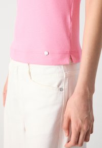 Close-up van een persoon die een roze geribbelde top en hoge witte broeken draagt, met een zichtbare zonnentattoo op de pols.