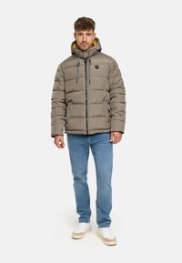 Pufferjakke i dempet grå med hette, har glidelås, quiltet design og kontrastfarget gul foring. Kombinert med lyseblå jeans og joggesko.