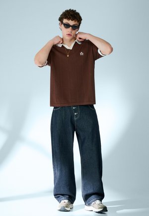 Junger Mann mit Sonnenbrille, braunem kurzärmeligen Strickshirt über weißem Kragen, weit geschnittenen dunklen Jeans und Turnschuhen, der den Kragen mit den Händen richtet.