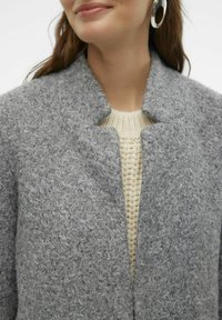 Grauer strukturierter Wollmantel ohne Kragen, kombiniert mit einem hellen cremefarbenen Strickpullover. Bemerkenswerte Details sind eine lockere Passform und weicher Stoff.