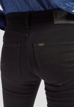 Nahaufnahme einer Person, die schwarze Lee-Jeans mit sichtbaren Gesäßtaschen, Gürtelschlaufen und einem in die Hose gesteckten blauen Denimhemd trägt.