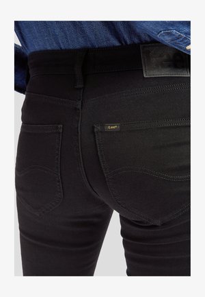 Primer plano de una persona que lleva vaqueros negros Lee con bolsillos traseros visibles, trabillas para cinturón y una camisa vaquera azul metida dentro.