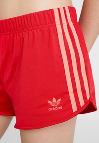 Červené sportovní šortky Adidas na boku a horní části stehna osoby, s třemi růžovými bočními pruhy a logem Adidas trefoil.