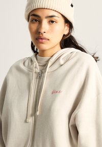 Beige Zip-Hoodie aus weichtexturiertem Material mit Kordelzügen und einem kleinen, rosa gestickten Logo auf der Brust. Getragen mit einem hellen Oberteil darunter.