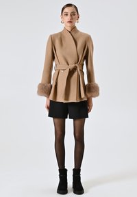 Cappotto avvolgente beige con polsini in pelliccia sintetica, cintura e due bottoni. Indossato sopra pantaloni corti neri e abbinato a stivali alla caviglia. Texture liscia.