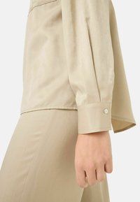 Blouse beige légère au toucher doux. Présente une coupe décontractée, un poignet boutonné et un éclat subtil. Fait partie d'une tenue assortie.