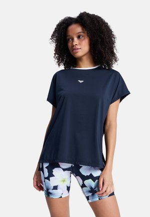 Vrouw met krullend haar draagt een loszittende marineblauwe korte mouwen top en zwarte fietsbroek met bloemmotief tegen een witte achtergrond.