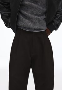 Sorte højttaljede bukser med læg kombineret med en grå striktrøje og en sort læderjakke med hænderne i lommerne.