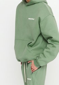 Persona che indossa una felpa con cappuccio e pantaloni della tuta dello stesso verde salvia con la scritta "MULTIPLY", mano nella tasca, su uno sfondo bianco semplice.