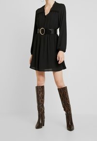 Svart långärmad klänning med V-ringning och spetsdetaljer, åtsnörd i midjan med ett blankt svart bälte med ett runt guldfäste. Bruna stövlar med ormskinnstryck som går till knäet.