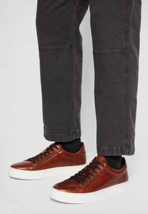 Brązowe skórzane trampki niskie z czarnymi sznurowadłami, noszone z czarnymi skarpetkami i ciemnoszarymi spodniami na jasnoszarym tle.