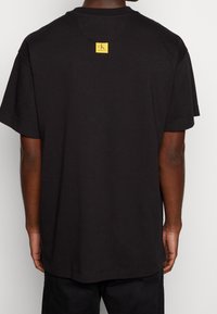 Schwarzes Baumwoll-T-Shirt mit einem gelben cK-Label am Halsausschnitt, locker sitzend und mit kurzen Ärmeln.