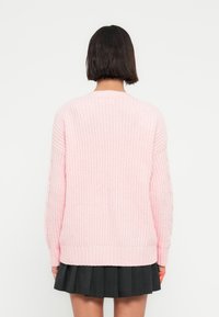 Ροζ πλεκτό πουλόβερ με υφασμένη υφή, μακριά μανίκια και ριμπ στο τελείωμα, συνδυασμένο με μαύρη ριγέ φούστα.