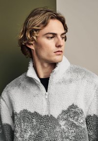 Polarowy półgolf w jasnoszarym kolorze z ciemnoszarymi wzorami. Posiada kołnierz z pół zamkiem i teksturowaną, puszystą powierzchnię. Miękki wygląd.