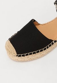 Svart kanvas espadrille med en yttersåle av jute-rep og en sydd kant, med en rund tå og design med åpen bak.