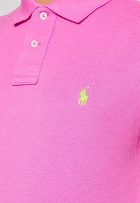 Ljusrosa polo-shirt i texturerad tyg, med krage, två knappar och en liten limegrön broderad logotyp.