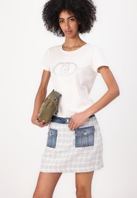 Hvit t-skjorte med metallisk trykkdesign, kombinert med en teksturert lyseblå skjørt med denimlommer. Modellen holder en olivengrønn veske.