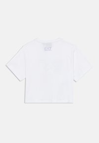 Baltas trumpomis rankovėmis marškinėlis su trumpintu dizainu, ant nugaros netoli kaklo išsiuvinėtas mažas levandų spalvos logotipas. Pagamintas iš minkštos medvilnės.