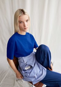 T-shirt en coton bleu associé à un haut style tablier rayé bleu, offrant une coupe décontractée et un détail de étiquette subtil, sur une personne assise.