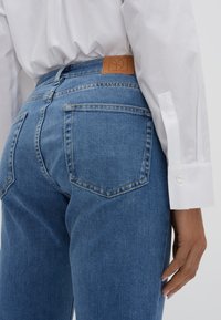 Jeans in denim blu a vita alta, con una toppa in pelle marrone testurizzata sul retro e tasche posteriori tradizionali.