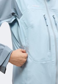 Χέρι που κλείνει την πλαϊνή τσέπη σε ανοιχτό μπλε μπουφάν Mammut με μπροστινές τσέπες στο στήθος και γκρι λεπτομέρειες στα μανίκια.