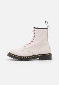White suede ankle boot with black accents, round toe, and a thick black sole featuring yellow stitching. Red and white laces add contrast.