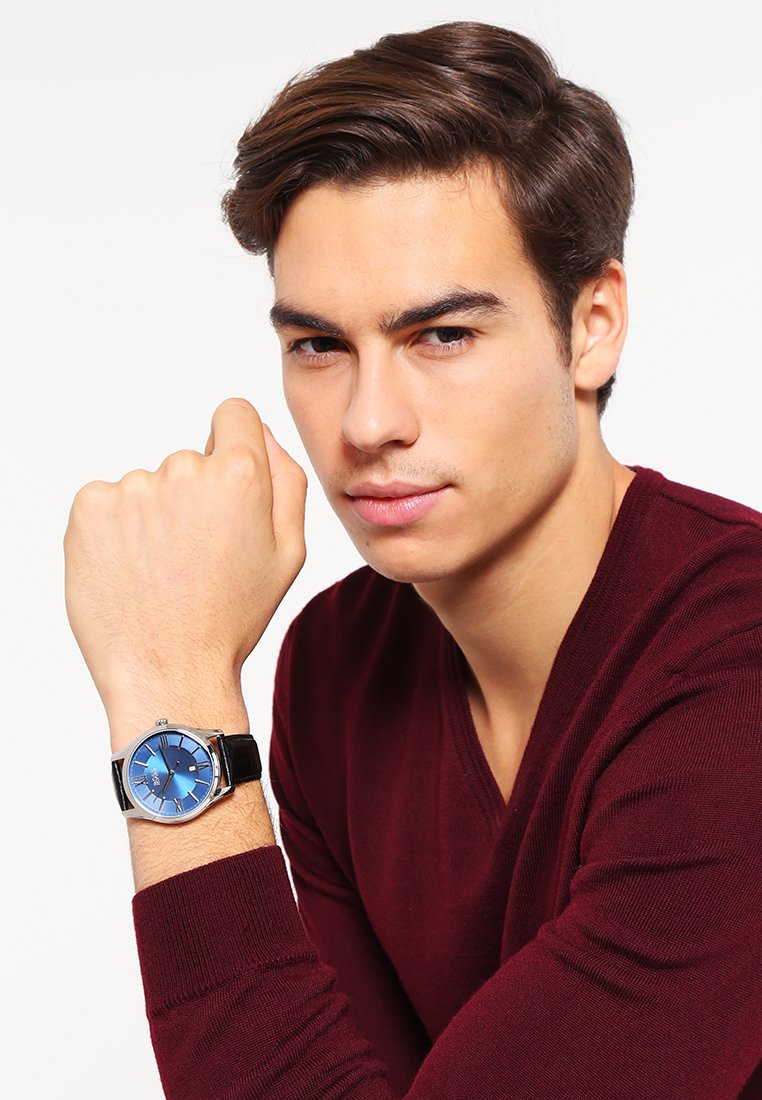 Montre pour homme avec un cadran bleu, des index argentés et un bracelet en cuir noir. Main reposant sur le poignet, portant un pull bordeaux.