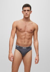 Graue Baumwoll-Slips mit einem schwarzen Logo-Bund, die eine enge Passform und glatte Textur bieten. Das Model zeigt eine stehende Pose mit nackter Haut.