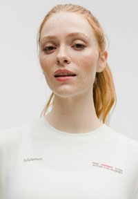 Rothaarige Frau mit Sommersprossen, die ein weißes Lululemon-Shirt mit dem Text "Finde den Fluss in der Reise" auf einem hellen Hintergrund trägt.