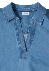 Chemise en denim bleu avec col et col en V profond, étiquette de marque visible "CECIL" au niveau du col.