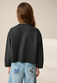 Schwarz beschnittenes Sweatshirt mit langen Ärmeln, kombiniert mit hellblauen Jeans, die blumen- und kirschgestickte Rückentaschen haben.