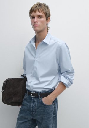 Ung mand iført lyseblå skjorte med knapper og op-rullede ærmer, cowboybukser, sort bælte, holder en mørkebrun ruskinds taske mod en ensfarvet hvid baggrund.