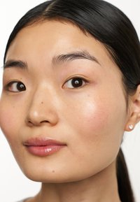Nahaufnahme eines Gesichts mit glatter Haut, natürlichem Make-up, leichtem Rouge und glänzenden, rosa Lippen. Das Haar ist dunkel und glänzend. Kleine Steckerohrringe sind sichtbar.
