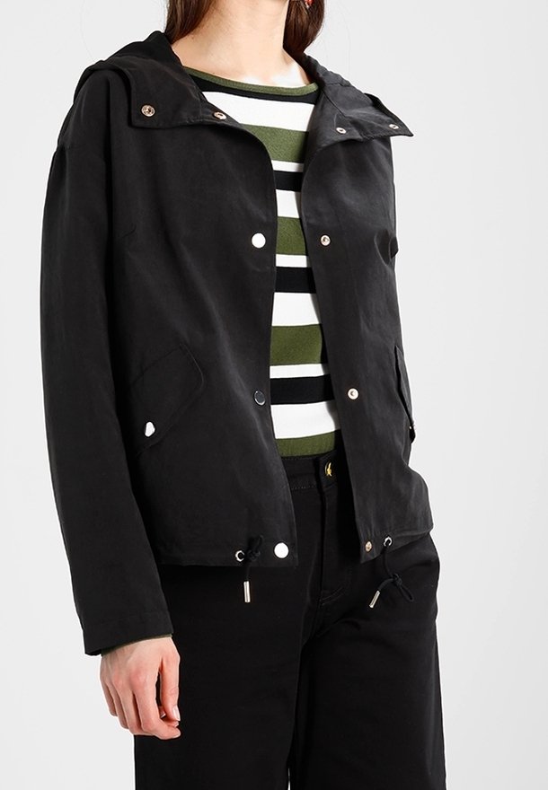 Black waterproof jacket with a hood, snap buttons, two front pockets, and drawstring hem. Worn over a green striped shirt and dark pants.