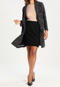 Manteau en tweed noir et gris avec des boutons, associé à un haut beige à côtes et une jupe noire. Le modèle porte des talons hauts noirs.
