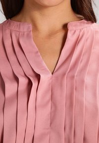 Blouse rose avec un col en V et un design plissé. Fabriquée en tissu lisse avec une texture douce. Col simple et coupe décontractée.