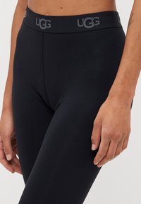 Juodi leggingai, pagaminti iš minkštos, tamprios medžiagos, su plačia juosmens dalimi, ant kurios yra pilkas UGG logotipas. Lygi tekstūra ir priglundantys dizainas.