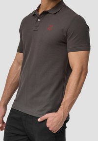 Polo en coton gris foncé avec un col, des manches courtes et un logo rouge brodé sur la poitrine, associé à un pantalon noir.