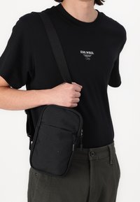 Czarna torebka na ramię wykonana z teksturowanego materiału, z zamkiem błyskawicznym, płaskim designem oraz regulowanym paskiem, trzymana na tle czarnej koszuli.