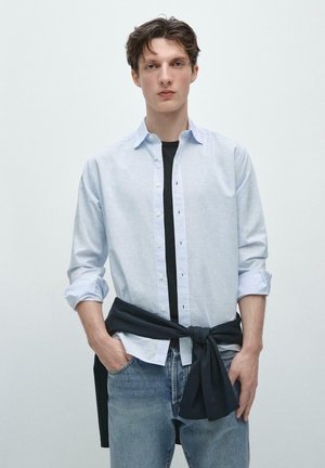 Ung mand iført en lyseblå skjorte over en sort t-shirt, blå jeans og en sort sweater bundet om livet, stående.