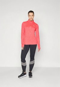 Crveni sportski top s dugim rukavima i poluzip ovratnikom, upareni s crnim tajicama sa sivim detaljima. Crne sportske tenisice upotpunjuju odjevnu kombinaciju.