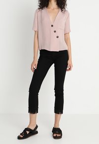 Kvinna i en ljusrosa kortärmad blus med knappar, avkortade svarta byxor med fransar samt svarta flätade sandaler.