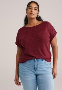 Bordeauxfarbener, strukturierter Pullover mit weitem Ausschnitt und aufgekrempelten Ärmeln, kombiniert mit hellblauen Jeans. Der Stoff zeigt ein dezentes Fischgrätmuster.