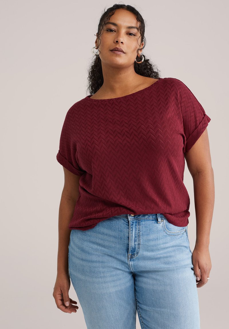 Bordeauxfarbener, strukturierter Pullover mit weitem Ausschnitt und aufgekrempelten Ärmeln, kombiniert mit hellblauen Jeans. Der Stoff zeigt ein dezentes Fischgrätmuster.