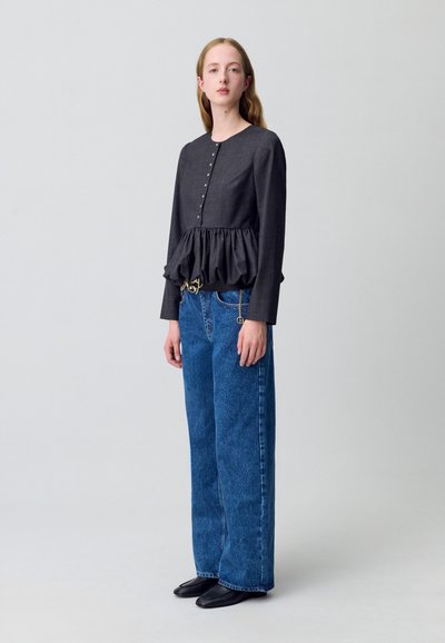 Una giovane donna sta in piedi indossando un top peplum scuro con bottoni e jeans blu a gamba larga, su uno sfondo leggero e uniforme.