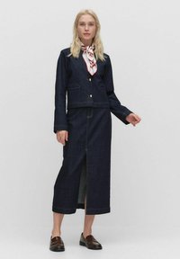 Ensemble en jean sombre avec une veste et une longue jupe, agrémenté de boutons dorés, de poches à rabat et d'une fente sur le côté. Le modèle porte un foulard à motifs et des mocassins marron.