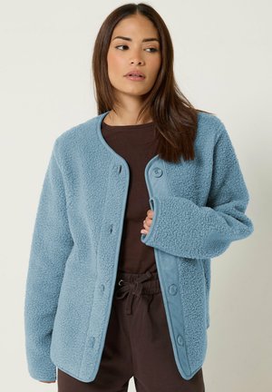 Femme aux longs cheveux bruns portant une veste légère bleu clair texturée et boutonnée, par-dessus une chemise et un pantalon marron foncé.