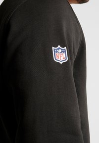 Felpa nera realizzata in tessuto testurizzato, con un logo NFL ricamato sulla manica sinistra con dettagli in rosso, bianco e blu.